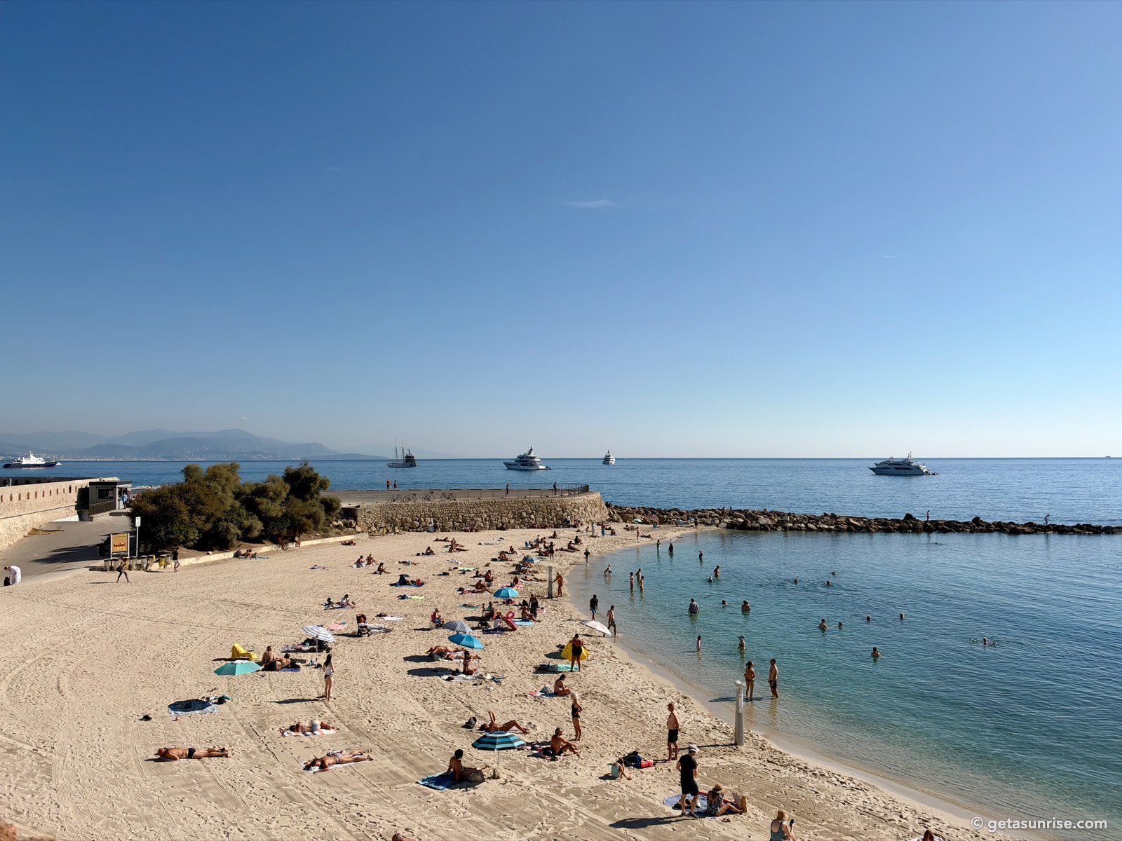 La Gravette beach in Antibes with swimmers, the harbour breakwater, yachts at anchor and the mountains around Nice on the horizon