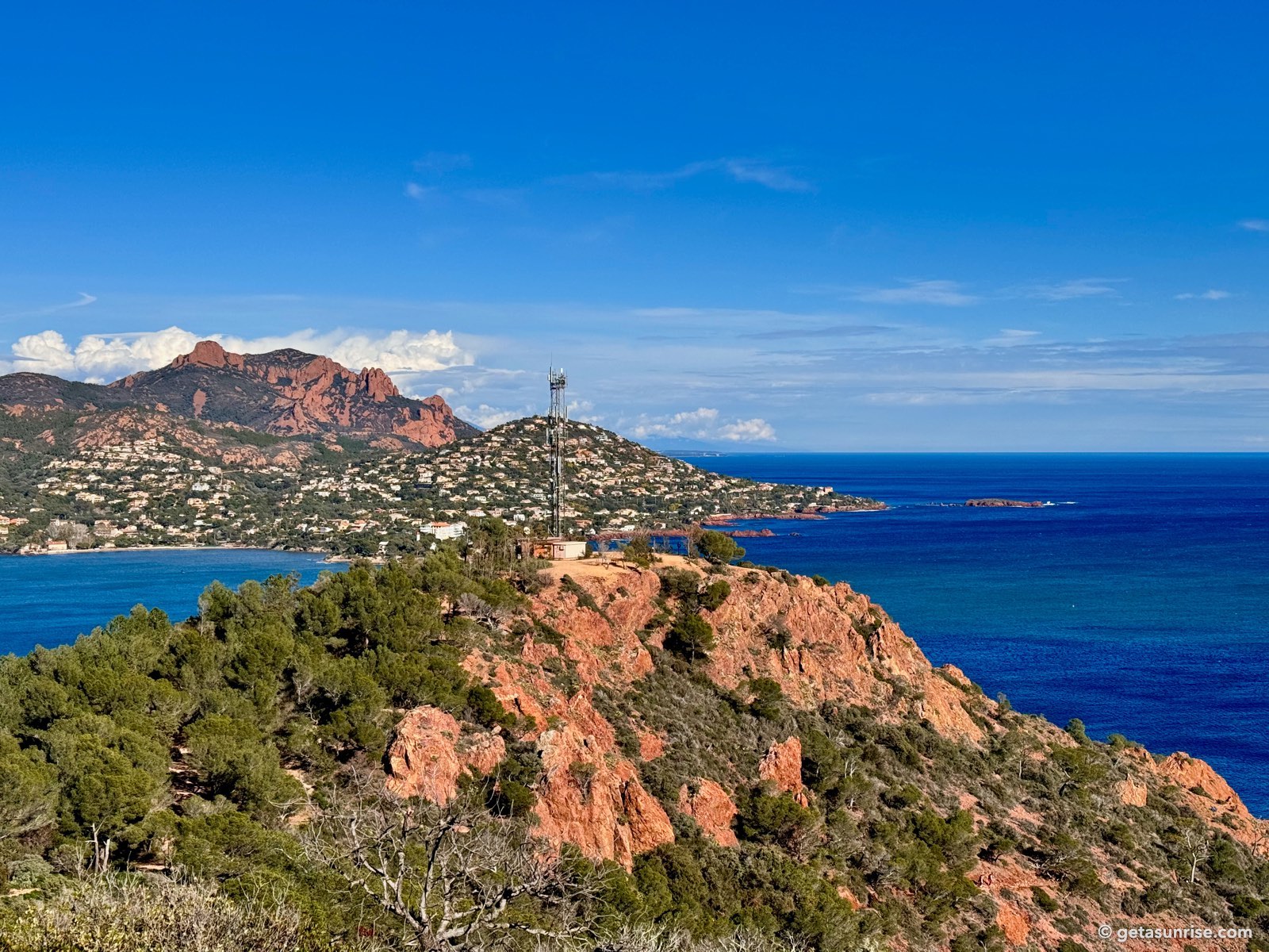 The bay of Agay seen from Cap Dramont, with the red rocks of the Esterel, villas on the hillside, a radio mast and the open sea on the right