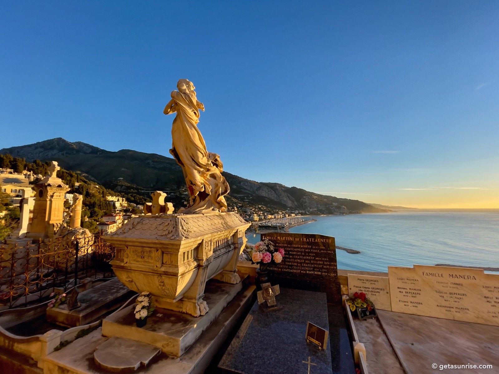 Statue on a grave at the Old Castle Cemetery in Menton, with the Garavan marina, the mountains and the coast towards Italy in the early morning light