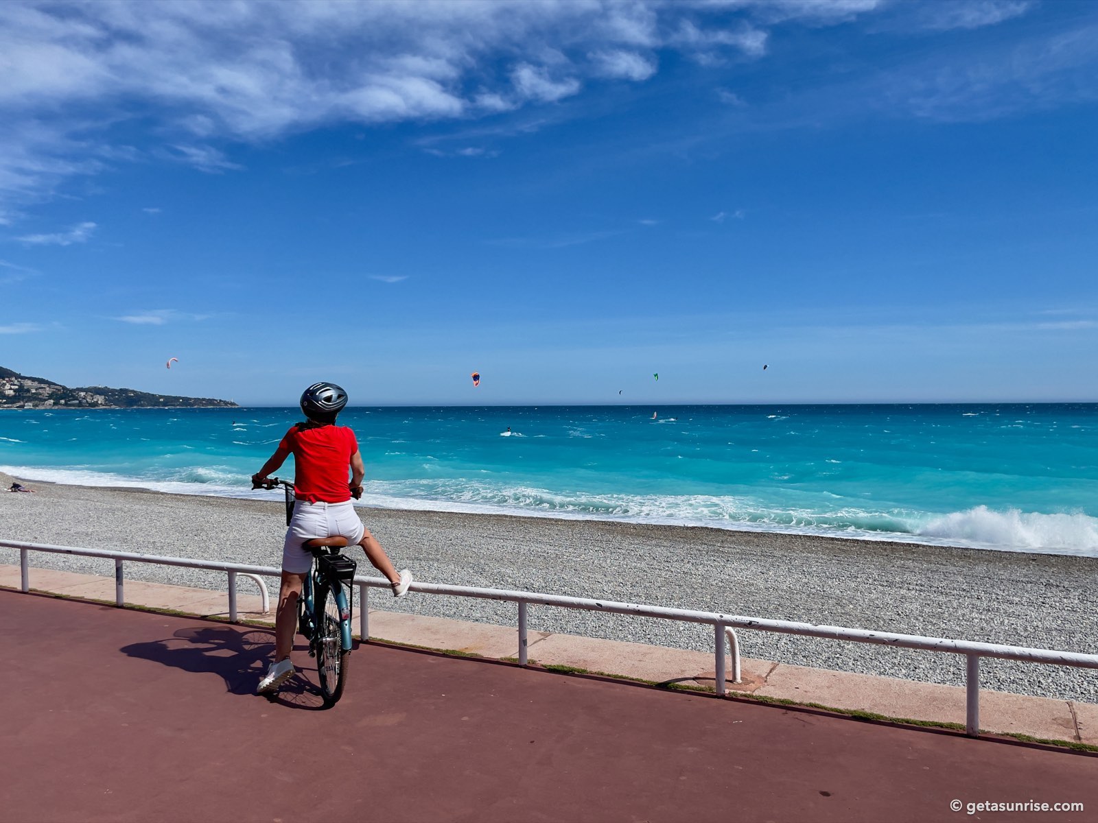 The Promenade des Anglais in Nice with a cyclist by the railing, the pebble beach, turquoise sea and the hill of Mont Boron on the left