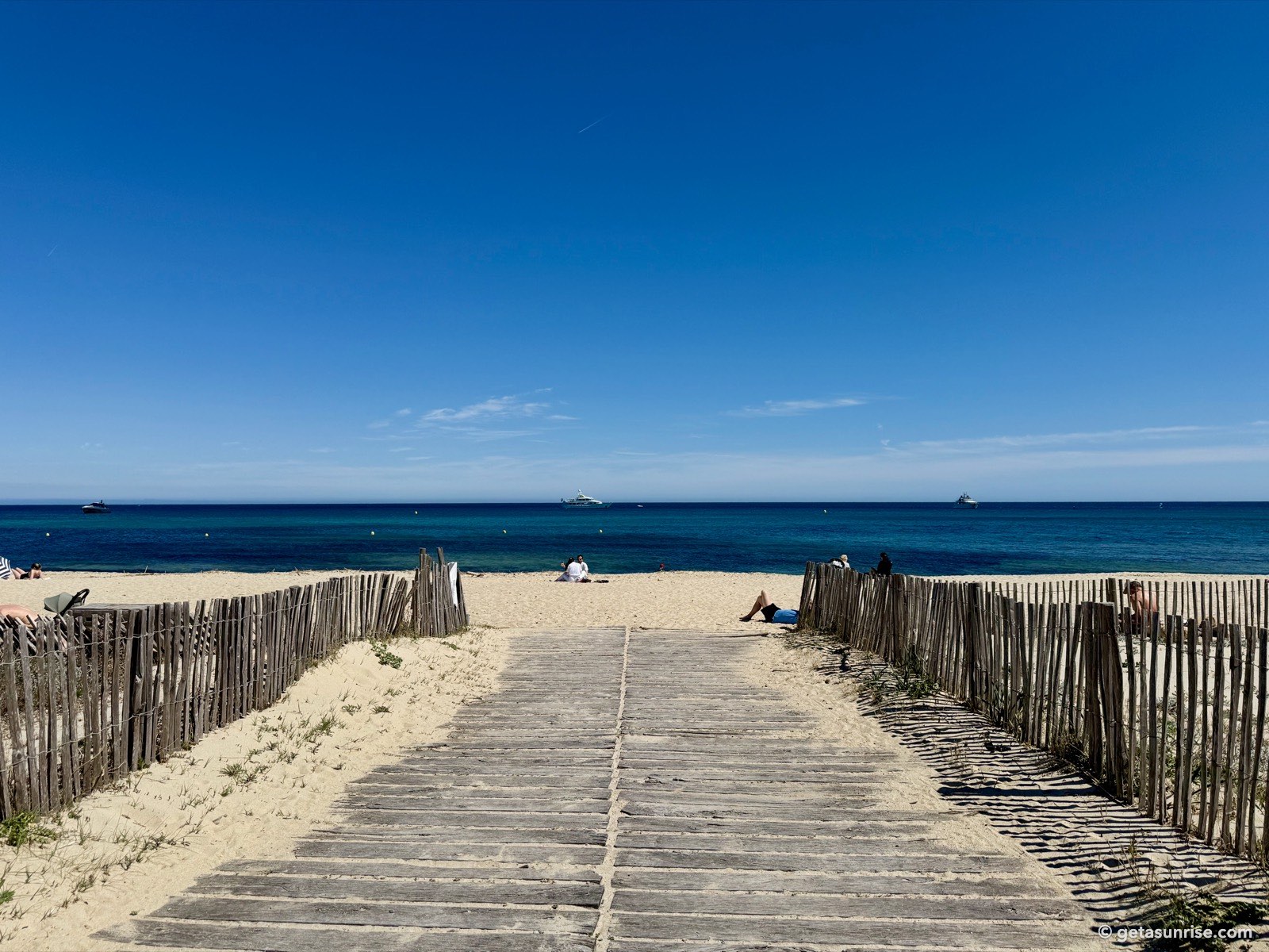 Wooden boardwalk between fences leading onto Pampelonne beach, with the open sea and a clear horizon